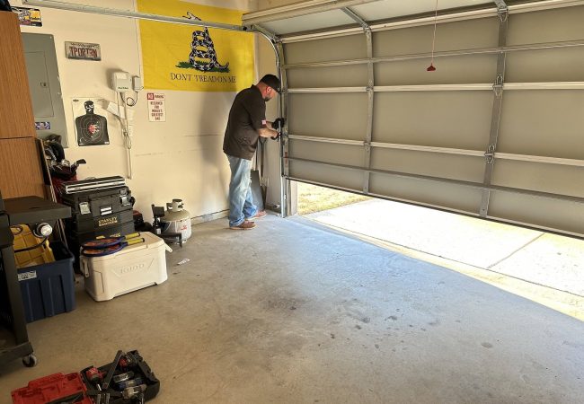 Person repairing a garage door in a residential garage, featuring tools and a "Don't Tread on Me" flag, illustrating emergency garage door repair services relevant to Fort Myers' unique climate challenges.