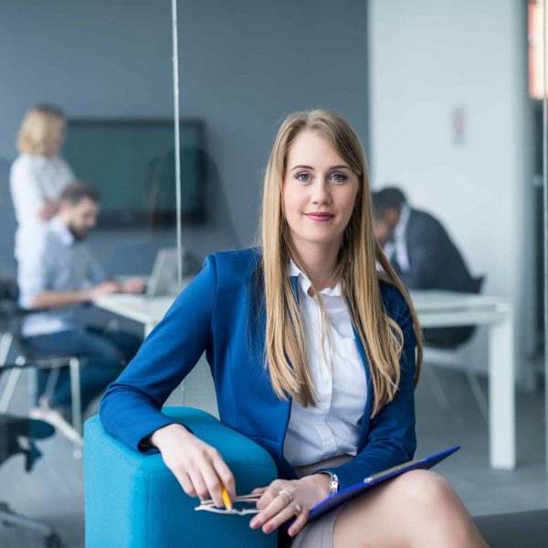 Businesswoman in a blue blazer sitting confidently in a modern office, holding a clipboard, with colleagues working in the background, representing professionalism and expertise in service delivery.