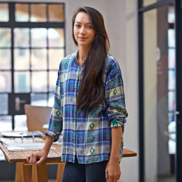 Young female designer in a modern office setting, wearing a patterned shirt, standing beside a wooden table with design materials.